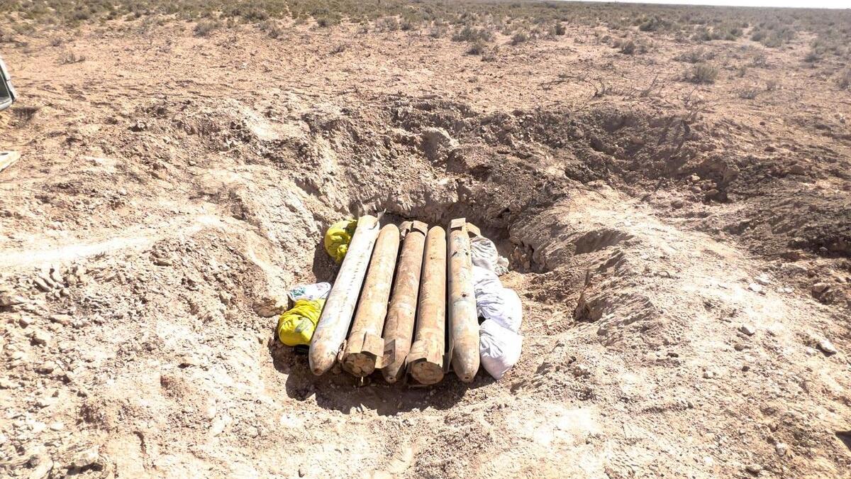Two field operators are shown in the midst of handling the ordnance. Both men are wearing light grey uniforms, caps, and distinct blue protective body armor vests. They are wearing heavy-duty gloves and are carefully passing a rusted projectile from one person standing on the ground to another standing on the bed of an orange utility truck. The truck features a cage-like barrier behind the cab and a red flag attached to a pole, serving as a safety warning signal.
