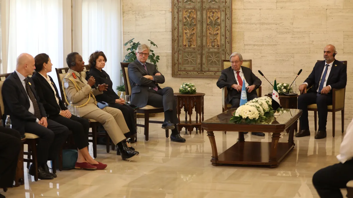 Delegates gather in a formal meeting room around a central table with floral arrangements and flags, as a uniformed speaker addresses the group.
