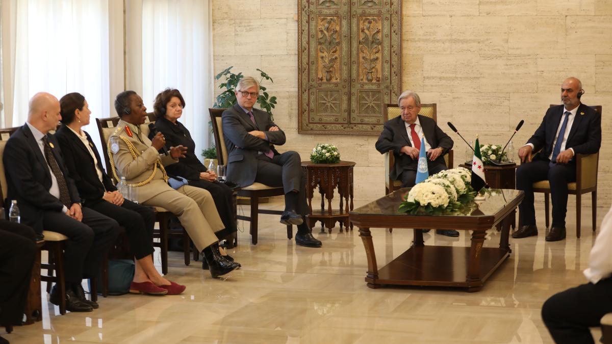 Delegates gather in a formal meeting room around a central table with floral arrangements and flags, as a uniformed speaker addresses the group.
