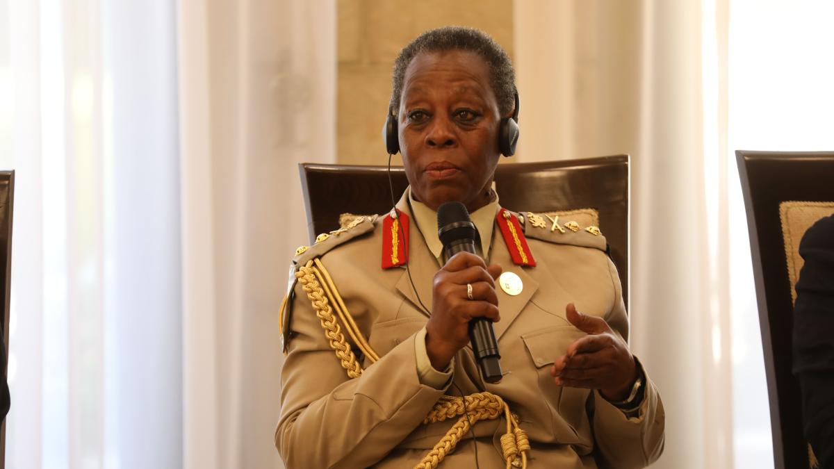 A uniformed official wearing a headset speaks into a handheld microphone while seated during a formal conference or diplomatic meeting.