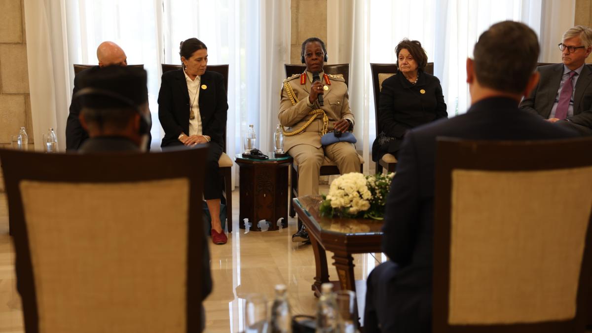 Participants sit in a formal meeting room as a uniformed official speaks into a microphone during a roundtable discussion, with attendees listening from both sides.