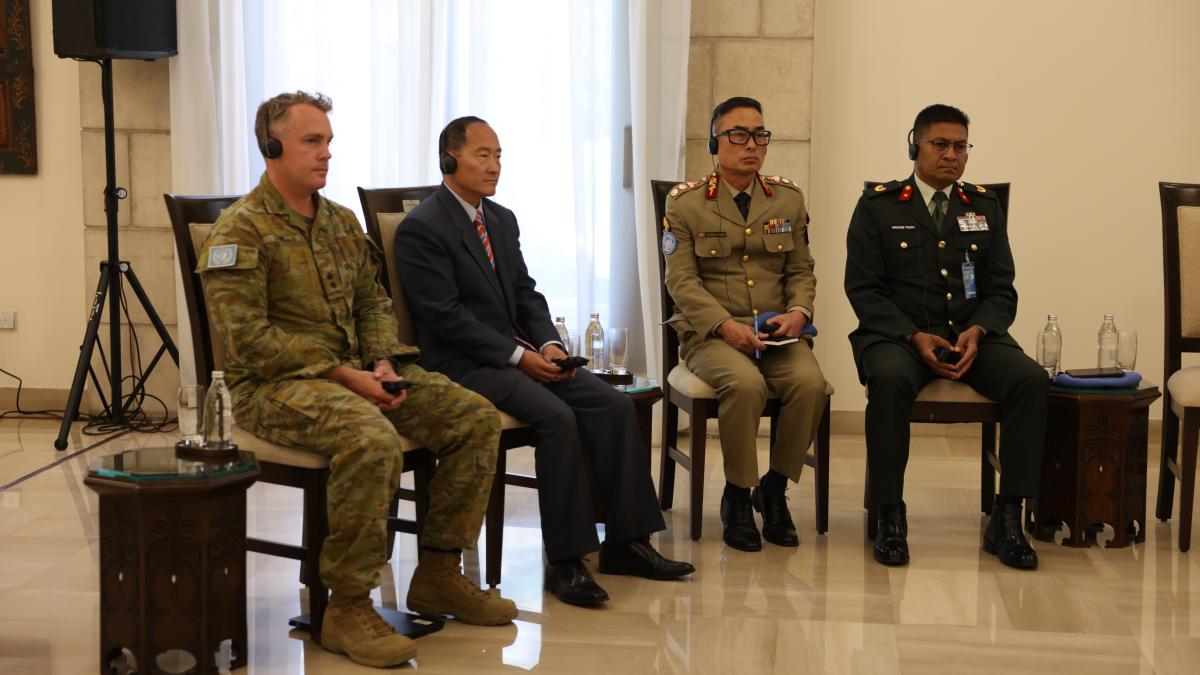 Four meeting participants wearing headsets, including military and civilian representatives, sit in a row during an international conference.