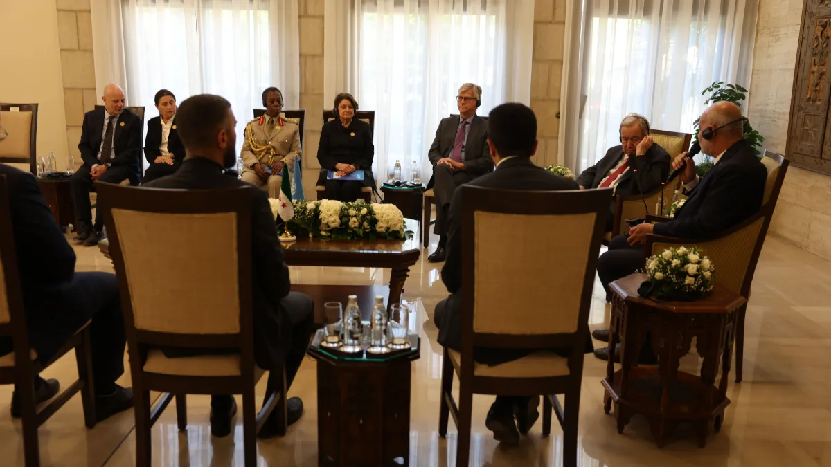 Delegates sit in a formal meeting arranged around a central table with floral displays, listening as a participant speaks into a microphone.