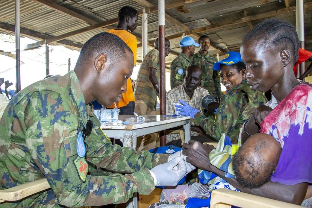 A person in camouflage uniform provides medical assistance to another person holding a baby at an outdoor clinic. Several people in uniforms and civilian clothing are seated at tables in the background under a corrugated metal roof.