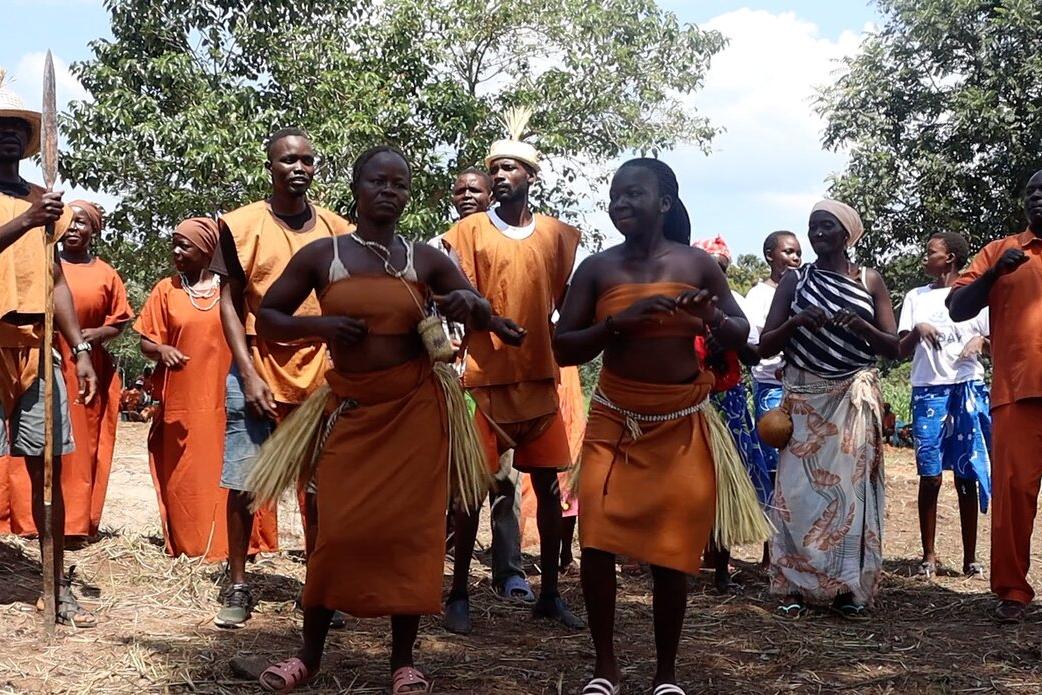 Group of people outdoors wearing traditional orange attire, some with grass skirts and necklaces, standing together with trees in the background.