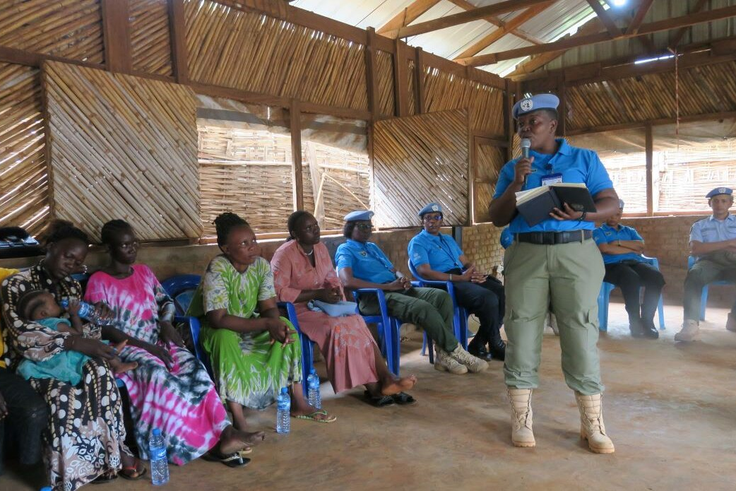 A UN peacekeeper in blue uniform stands and speaks with a notebook and microphone during a community meeting inside a wooden structure, while others sit on blue chairs listening