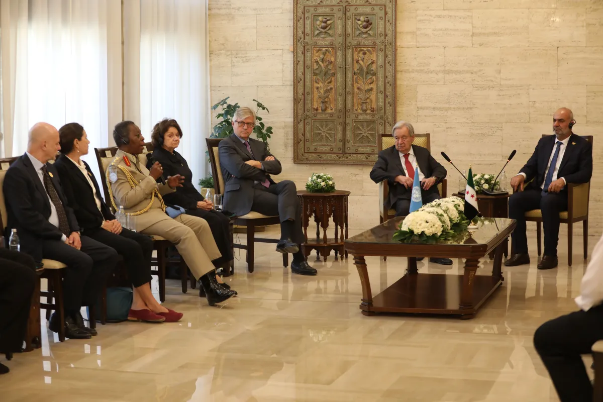 Delegates gather in a formal meeting room around a central table with floral arrangements and flags, as a uniformed speaker addresses the group.