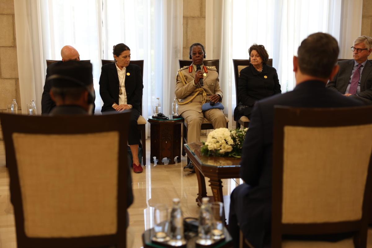 Participants sit in a formal meeting room as a uniformed official speaks into a microphone during a roundtable discussion, with attendees listening from both sides.