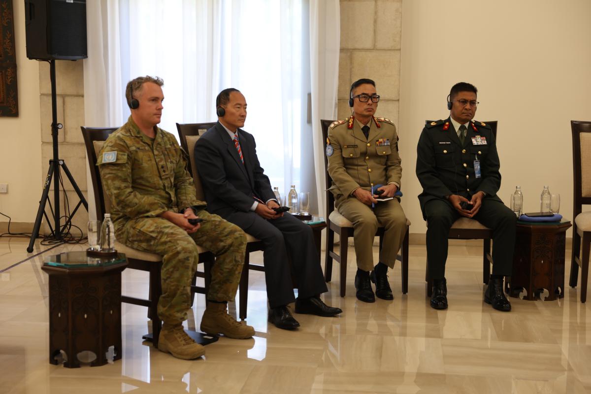 Four meeting participants wearing headsets, including military and civilian representatives, sit in a row during an international conference.