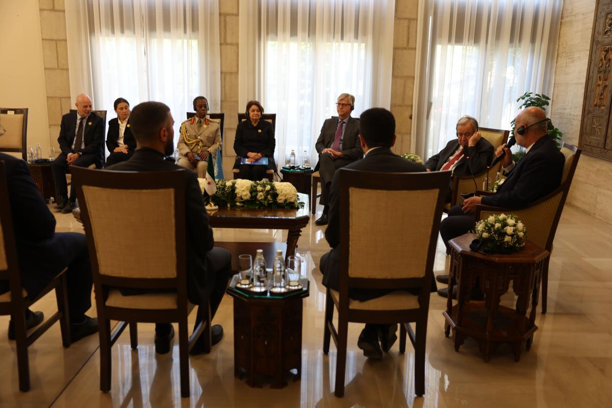 Delegates sit in a formal meeting arranged around a central table with floral displays, listening as a participant speaks into a microphone.