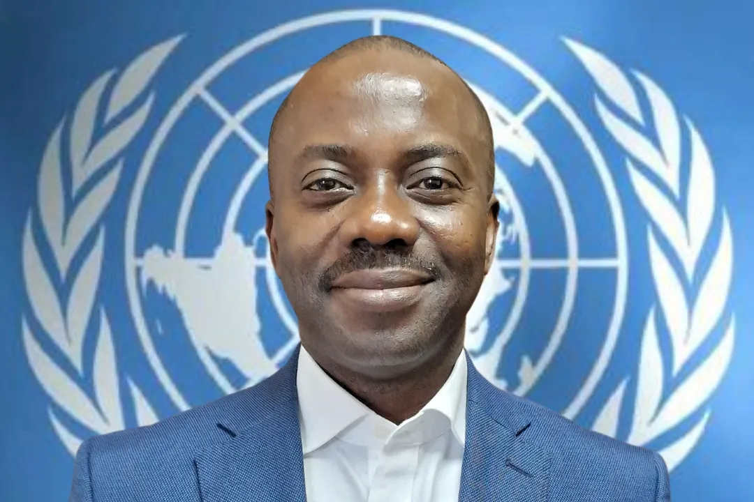 A headshot of a man in a suit stands in front of the UN logo