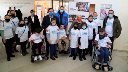 Group of people, including children and adults, standing indoors wearing white T-shirts with blue text promoting equality and freedom. Two individuals are in wheelchairs. Banners and posters are visible in the background.
